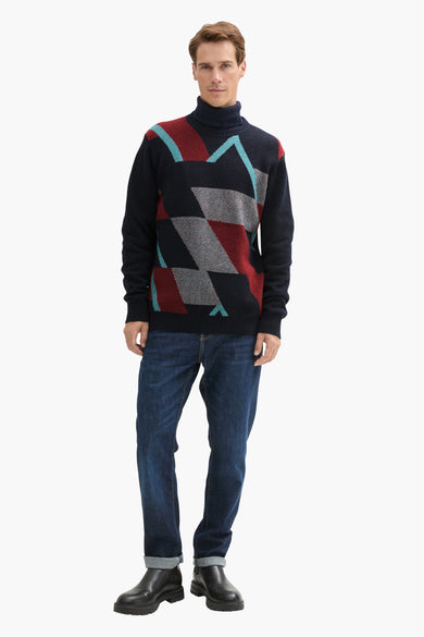 Blauwe trui met ronde hals van TOM TAILOR, met geometrische patronen in rood, grijs en turquoise, gecombineerd met jeans en zwarte laarzen.