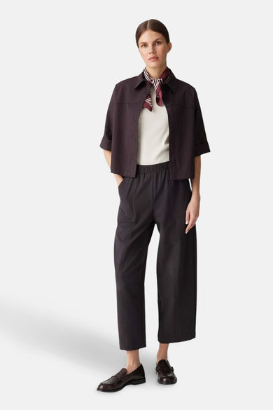 Pantalon brun d'OPUS, avec une ceinture élastique et des jambes larges, porté avec un chemisier blanc et un foulard rayé.