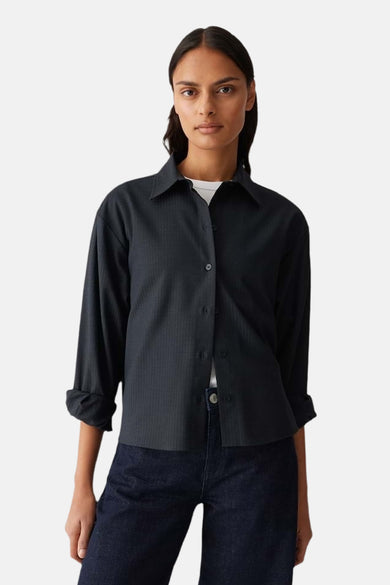 Vrouw draagt zwarte blouse met lange mouwen en donkere jeans, staand tegen een witte achtergrond.