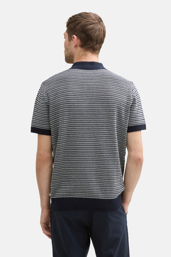 Achterkant van een man in een zwart en wit poloshirt en donkerblauwe broek, met zijn handen in zijn zakken.