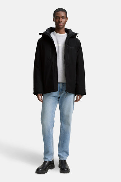 Manteau court noir de TOM TAILOR, avec capuche, associé à un pull gris et des jeans bleus.