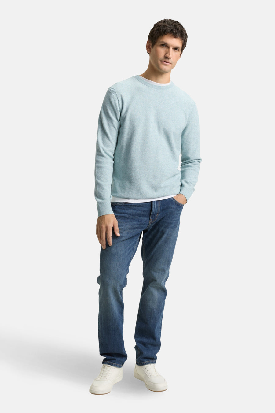 Man in een lichtblauwe trui, jeans en witte sneakers.