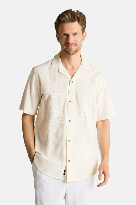 Chemise écru à manches courtes de TOM TAILOR, avec feuilles brodées et col cubain, portée avec un pantalon blanc et des sandales.