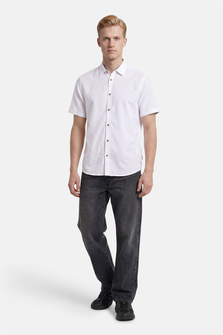 Chemise blanche à manches courtes de TOM TAILOR, avec des boutons marron, portée par un homme avec un jean foncé et des baskets noires.