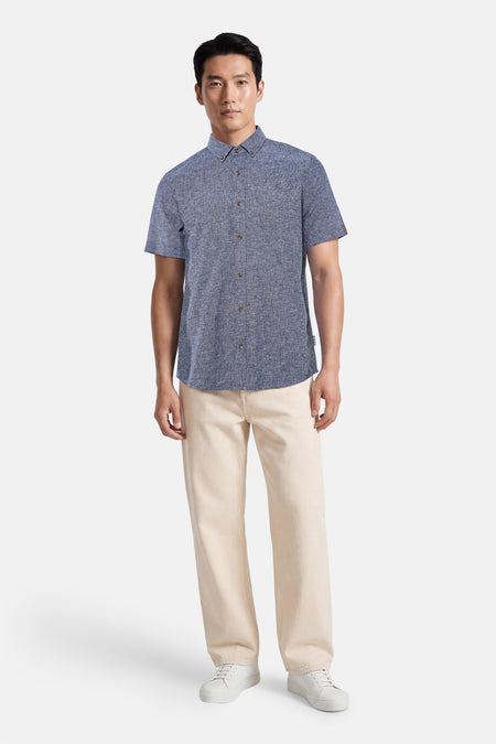 Chemise bleue à manches courtes de TOM TAILOR, avec tissu texturé, col boutonné et boutons marron, portée par un homme avec un pantalon beige clair et des baskets blanches.