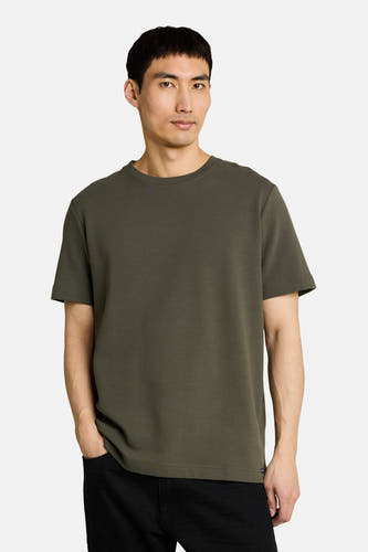 Kaki T-shirt met korte mouwen van TOM TAILOR met een ronde hals en fijne ribstructuur, gedragen door een man met handen in zwarte broekzakken.