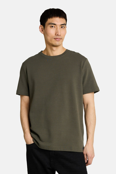Kaki T-shirt met korte mouwen van TOM TAILOR met een ronde hals en fijne ribstructuur, gedragen door een man met handen in zwarte broekzakken.