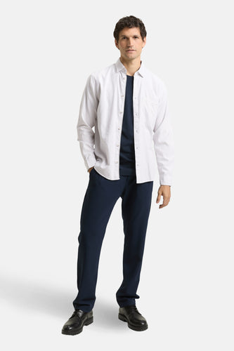 Een man in een blauw shirt en een open wit overhemd, met een hand in zijn zak, draagt donkerblauwe broeken en zwarte schoenen.