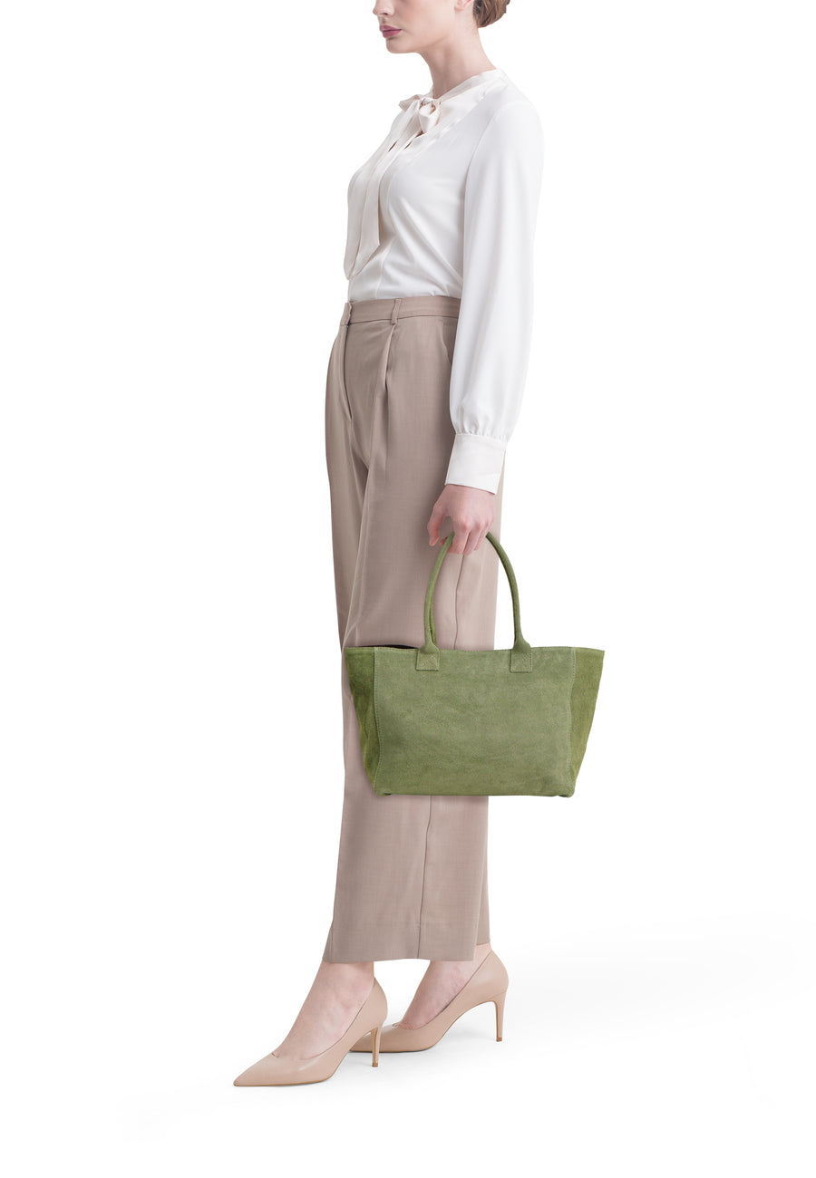 Khakikleurige suède shoppertas van Access®, gedragen door een vrouw in een witte blouse en beige broek.