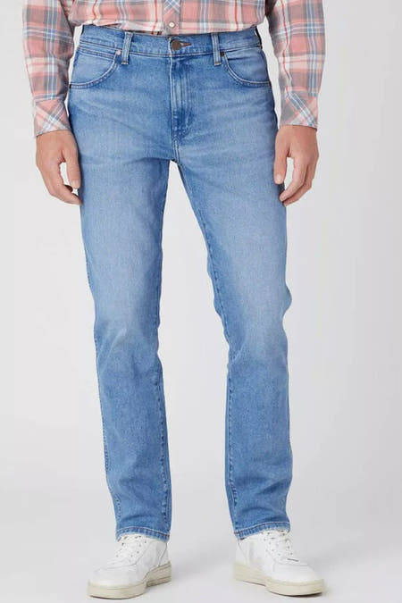 Lichtblauwe tapered jeans van Wrangler, gecombineerd met een roze geruit overhemd en witte sneakers.
