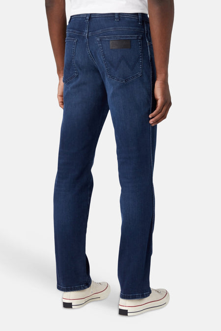 Achteraanzicht van donkerblauwe straight jeans van Wrangler® met zwarte patch en 'W'-stiksels op de achterzakken, gedragen met witte sneakers.