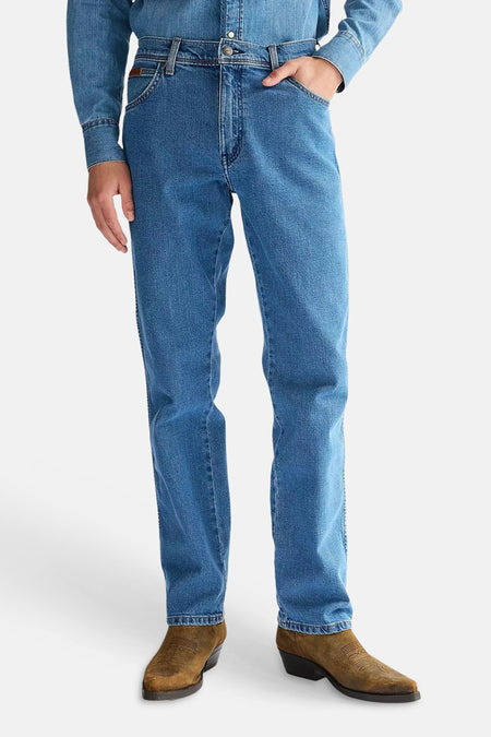 Rechte jeans in middenblauw denim van Wrangler, gedragen met een denim overhemd en bruine cowboylaarzen.