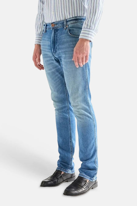 Lichtblauwe straight jeans van Wrangler, gedragen door een man met een gestreept overhemd en zwarte instappers.