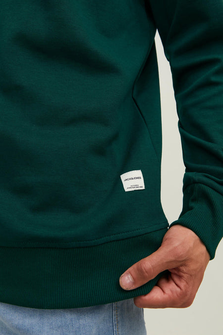 Donkergroene ronde hals sweater van ORIGINALS BY JACK & JONES met wit label op borst, ribboorden aan mouw en zoom, close-up van model in jeans.