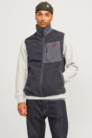Grijze bodywarmer van Royal Denim Divison, met fleece textuur en een grijze sweater eronder, gecombineerd met donkere jeans en een groene muts.
