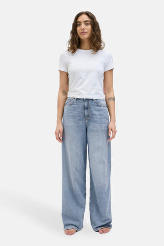 Lichtblauwe, wijde jeans van JJXX, gedragen met een wit T-shirt en blote voeten.
