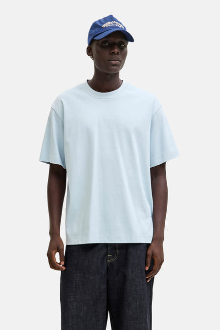 T-shirt bleu à manches courtes de CORE BY JACK & JONES, porté avec un chapeau bleu et des jeans larges, avec des baskets blanches.