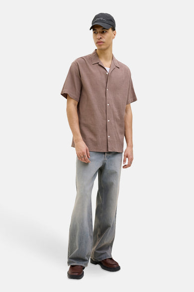 Chemise brune à manches courtes d'ORIGINALS par JACK & JONES, avec col boutonné, portée avec des jeans et un chapeau.