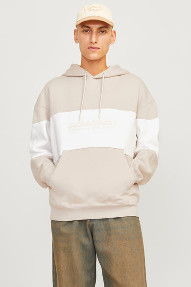 Man met beige hoodie en pet, denim broek, tegen een witte achtergrond.
