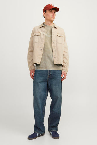 Een man in een beige jas, groen T-shirt, wijde jeans en een rode pet.