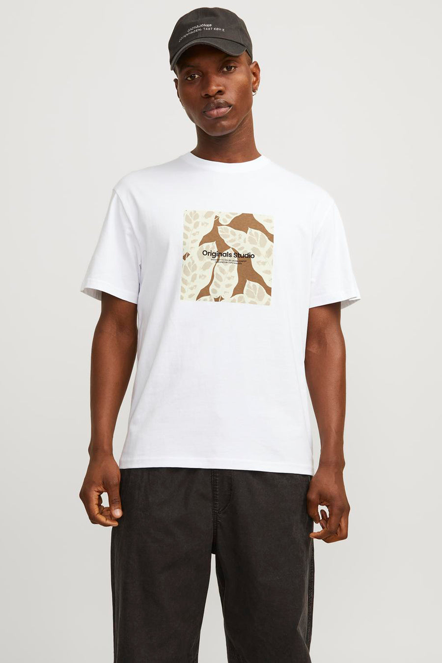 Een man met een donkerblauwe pet draagt een wit T-shirt met een bruine print en een donkergrijze broek.