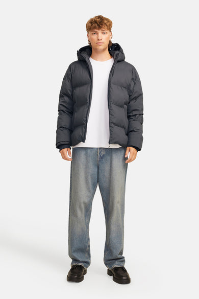 Zwarte regenjas van CORE BY JACK & JONES, gedragen met een wit T-shirt en wijde jeans, op een witte achtergrond.
