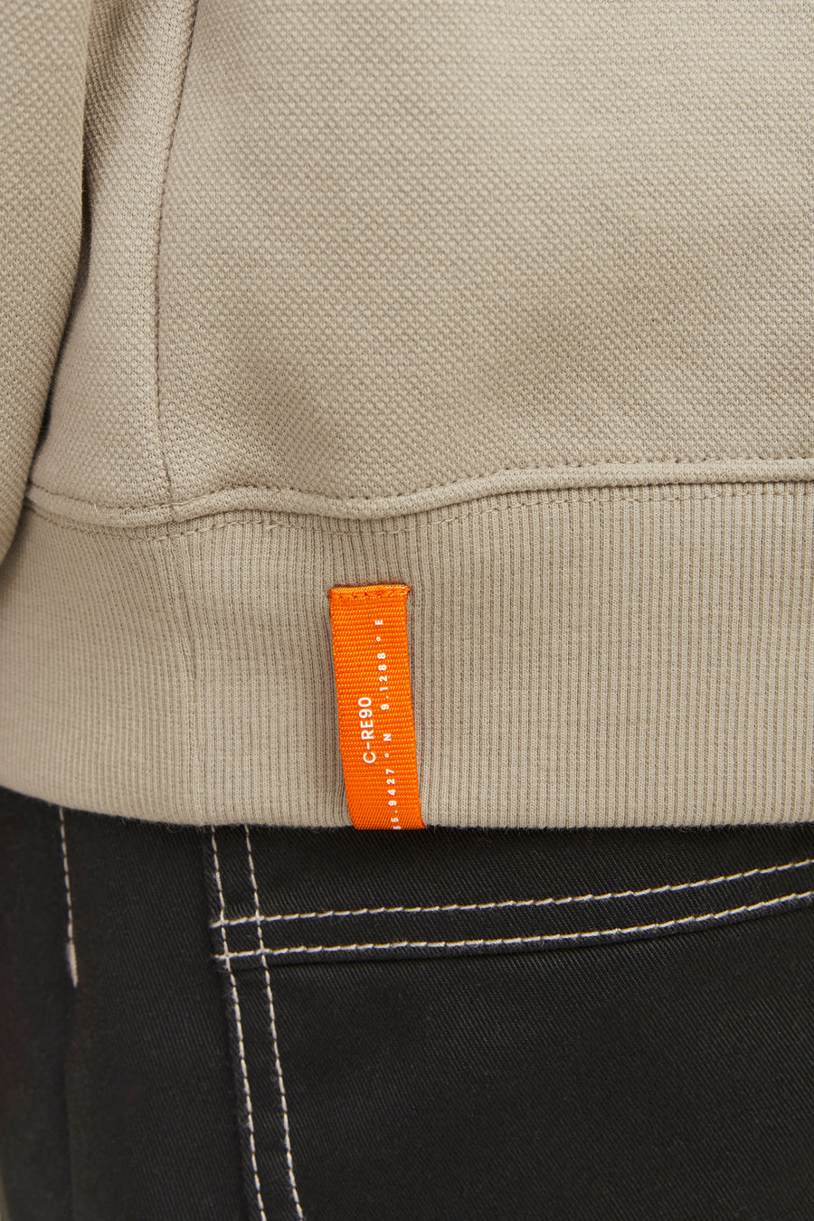 Beige trui met oranje label, zwarte broek met witte stiksels.
