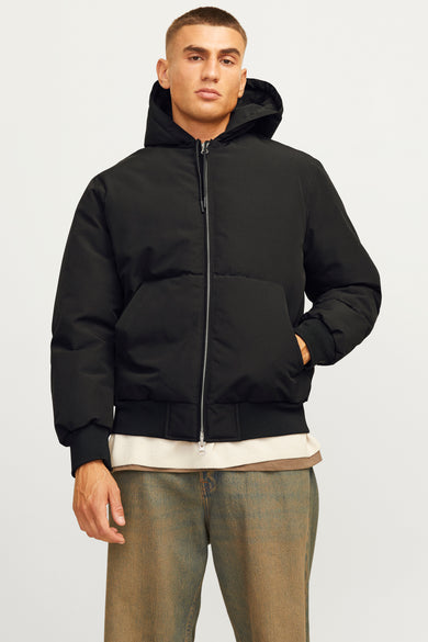 Zwarte korte jas van CORE BY JACK & JONES, met capuchon en zilveren rits, gedragen met een beige shirt en jeans.
