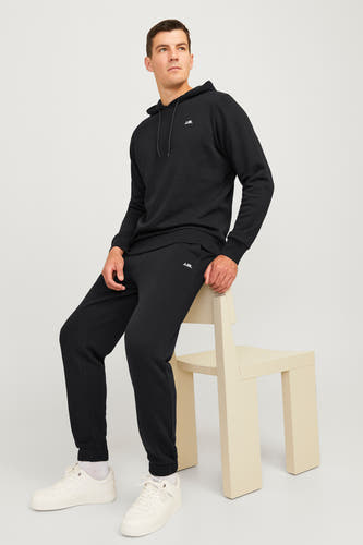 Zwarte jogging van JJREBEL, gedragen door een man met witte sneakers en sokken, zittend op een beige stoel.
