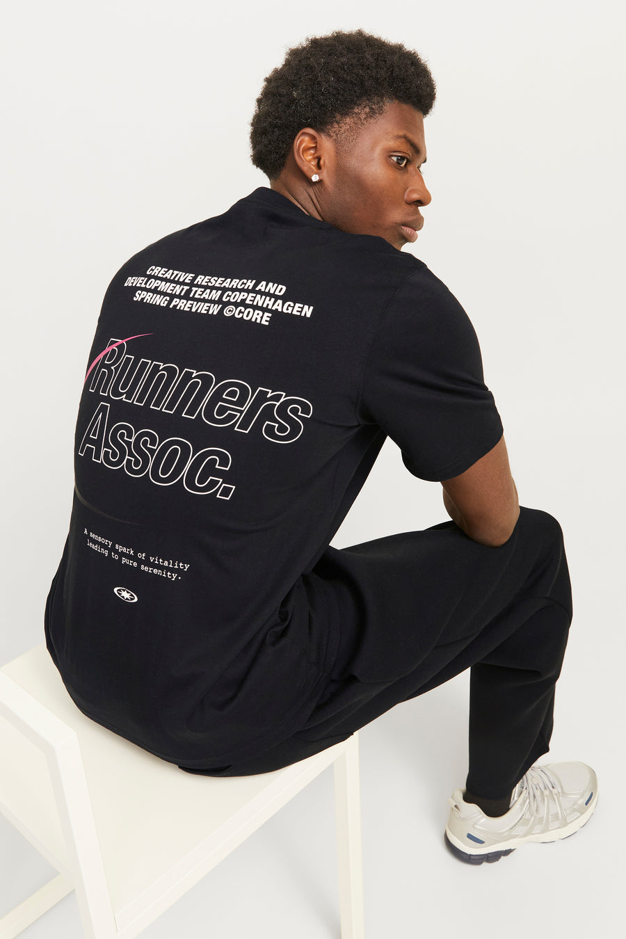 Zwart T-shirt met korte mouwen van CORE by JACK & JONES, met witte tekstprint op de achterkant, gedragen met een zwarte broek en zilverkleurige sneakers.
