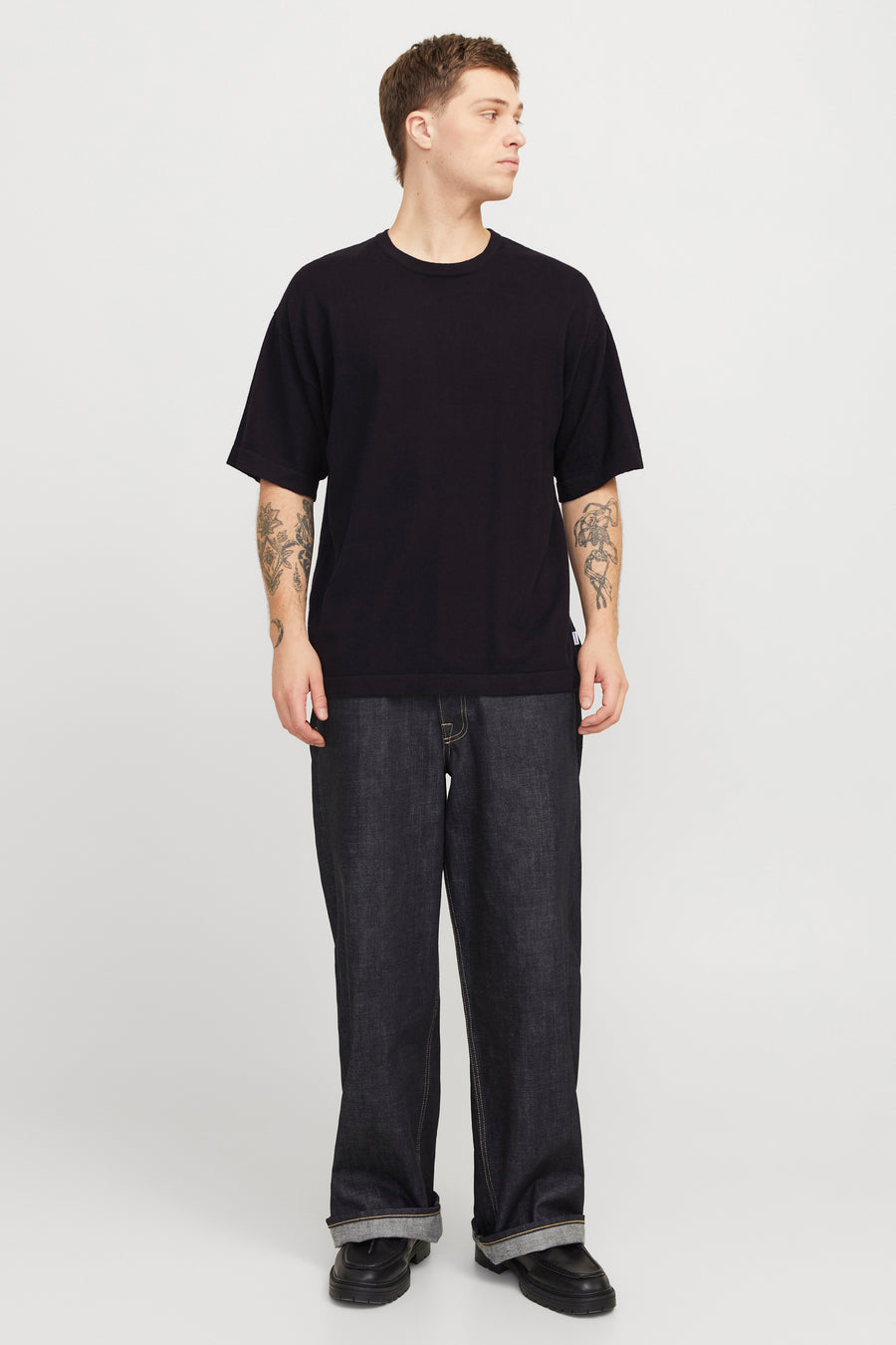 Zwart T-shirt met korte mouwen van PREMIUM by JACK & JONES, met tatoeages op de armen, gecombineerd met donkerblauwe jeans en zwarte schoenen.
