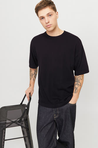 Zwart T-shirt met korte mouwen van PREMIUM by JACK & JONES, gedragen met donkere jeans en leunend op een metalen stoel.
