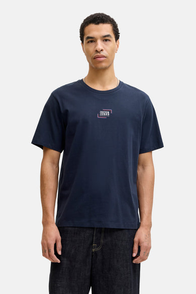 T-shirt bleu à manches courtes de CORE by JACK & JONES, avec un logo sur la poitrine, porté avec des jeans larges et des chaussures marrons.