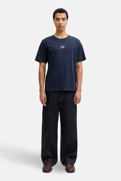 Blauw T-shirt met korte mouwen van CORE by JACK & JONES, met een logo op de borst, gedragen met wijde jeans en bruine schoenen.
