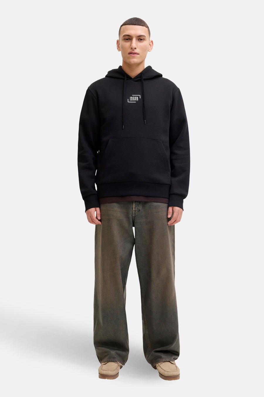 Man met zwarte hoodie, ruime jeans en suède mocassins op witte achtergrond.
