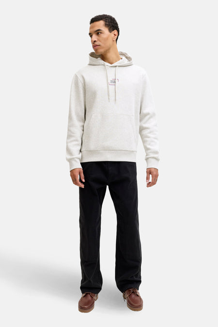 Een man in een grijze hoodie, zwarte broek en bruine schoenen, staand tegen een witte achtergrond.