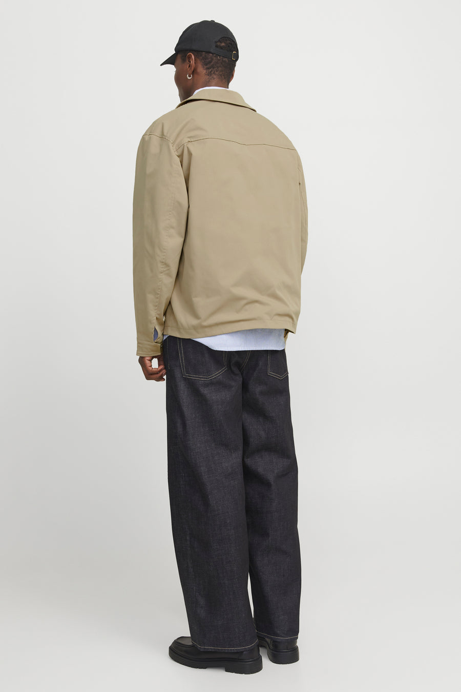 Persoon van achteren in beige jas, blauw gestreept overhemd, wijde donkere jeans en zwarte schoenen, met zwarte pet.