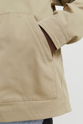 Manteau court beige d'ORIGINALS BY JACK & JONES, avec une main dans la poche et un détail d'un vêtement rayé.