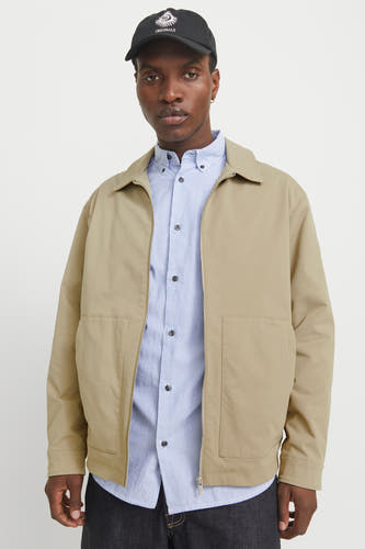 Manteau beige court d'ORIGINALS BY JACK & JONES, avec un chemisier rayé bleu et une casquette noire.
