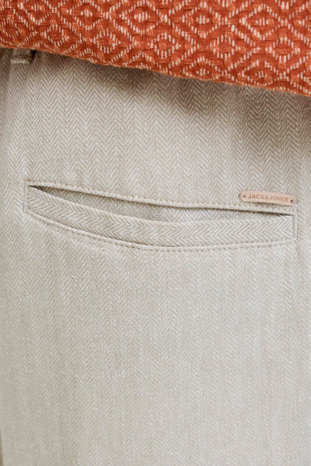 Close-up van een beige short van JACK & JONES JEANS INTELLIGENCE met visgraatpatroon, een achterzak en bruin merklabel, gestyled met een oranje gebreide top.