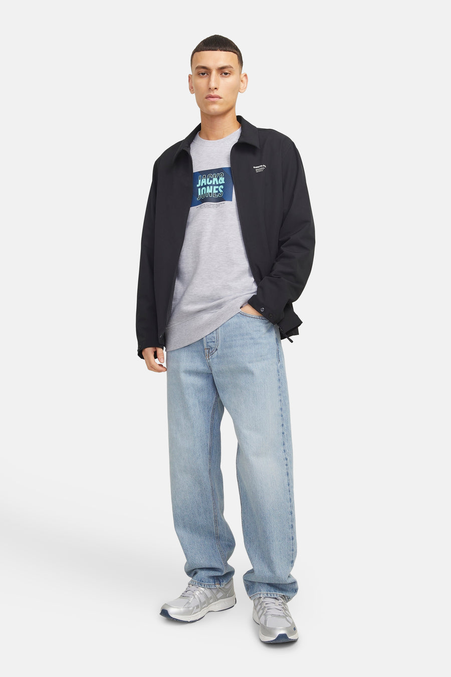 Pull-over gris à col rond de CORE by JACK & JONES, avec un logo sur la poitrine, porté avec une veste noire et des jeans larges.