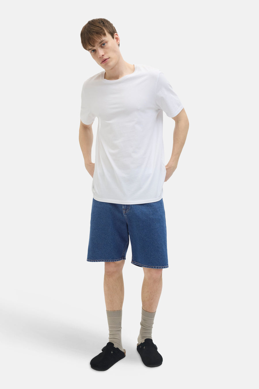 Jeansshort van JJREBEL, in medium blauw denim, gedragen met een wit T-shirt, grijze sokken en zwarte muiltjes.
