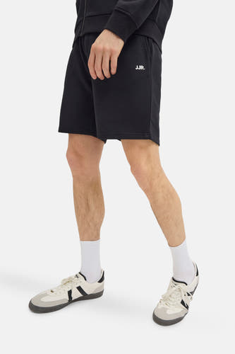 Short noir de JJREBEL, avec une impression de logo blanc, porté avec des chaussettes blanches et des chaussures crème.