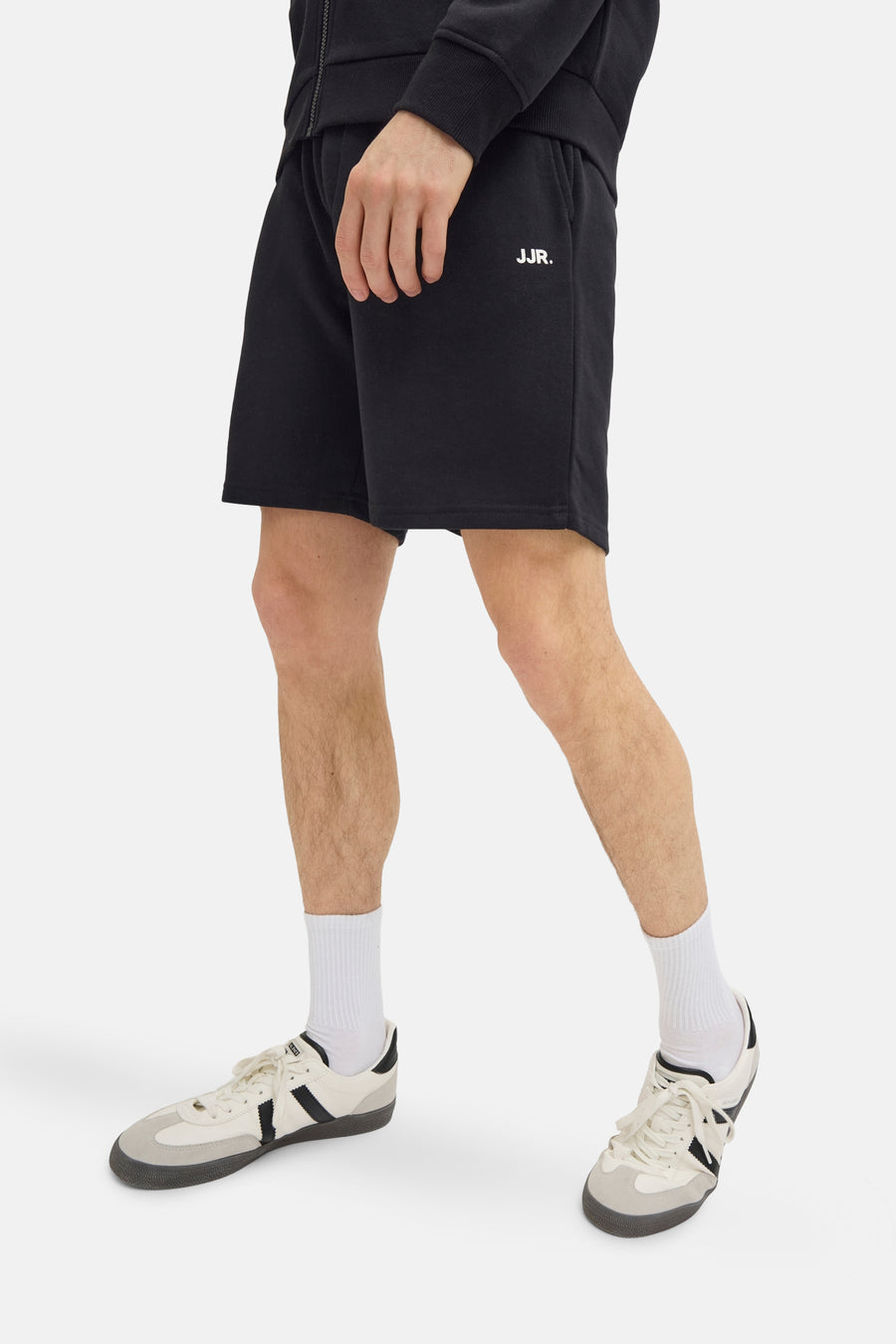 Short noir de JJREBEL, avec une impression de logo blanc, porté avec des chaussettes blanches et des chaussures crème.