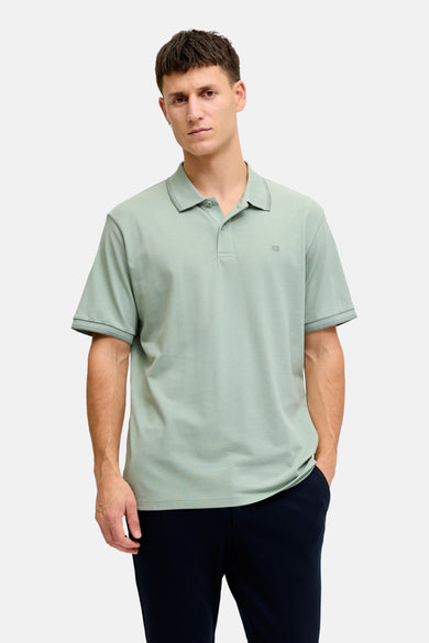 Polo verte JJREBEL de couleur verte à manches courtes, col côtelé et un petit logo 'R' sur la poitrine, porté avec un pantalon bleu foncé et des mocassins marron.