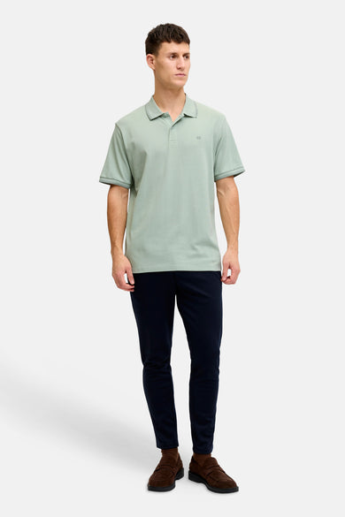 Groene polo van JJREBEL met korte mouwen, ribkraag en een klein 'R' logo op de borst, gedragen met donkerblauwe broek en bruine loafers.