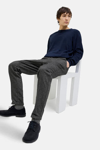 Grijze chino van JACK & JONES JEANS INTELLIGENCE, gedragen met een donkerblauwe trui en zwarte schoenen, zittend op een witte stoel.