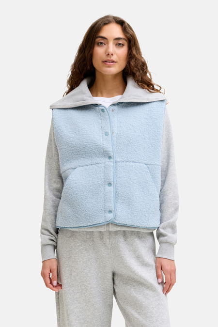 Blauwe bodywarmer van JJXX, met knoopsluiting en fleece textuur, gedragen over een grijze sweater en joggingbroek.
