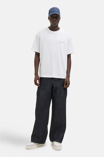 Wit T-shirt met korte mouwen van ORIGINALS BY JACK & JONES, met een kleine tekstprint en gedragen met een donkerblauwe pet en wijde jeans.
