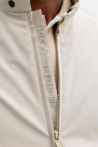 Beige korte jas van PREMIUM by JACK & JONES, met ritssluiting, verticale JACK & JONES PREMIUM print en zwarte drukknoop, over een lichtbruin poloshirt.